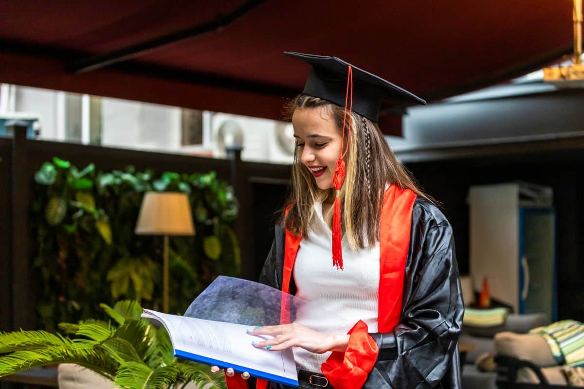 Jovem vestindo beca de formatura e lendo um livro, representando o investimento em educação superior.