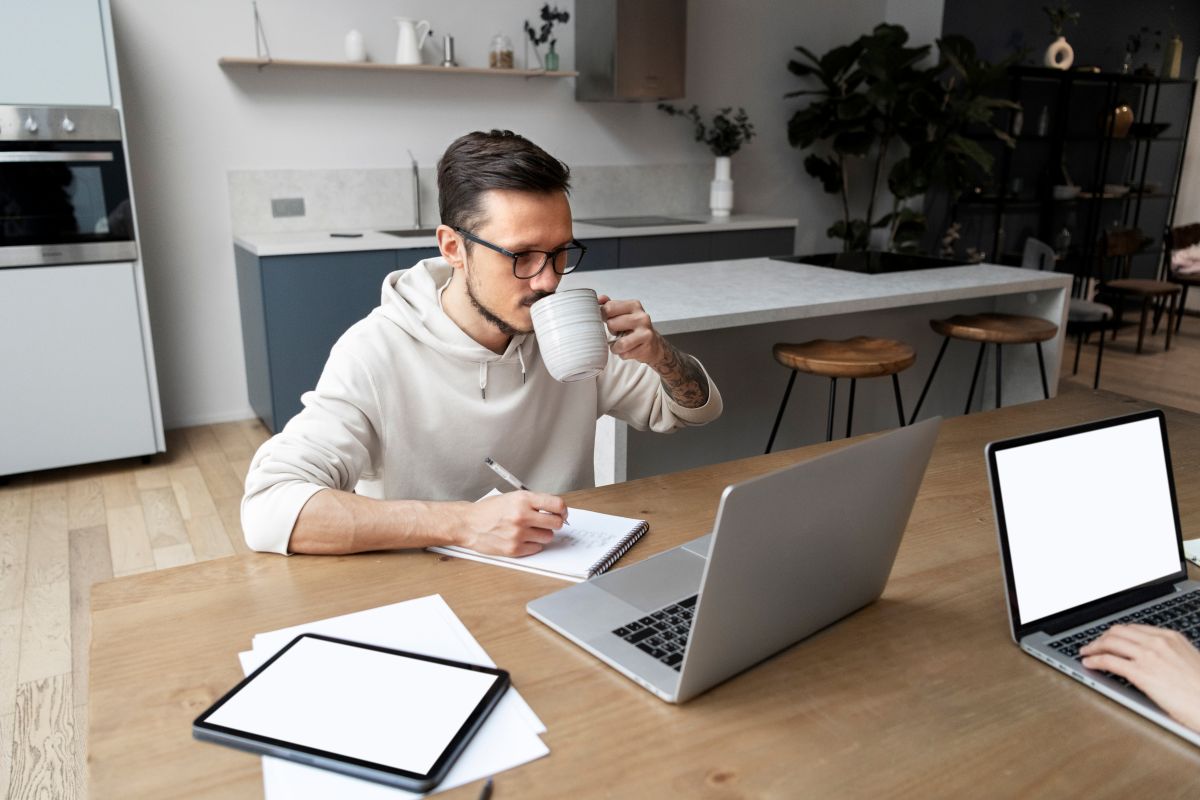 Um homem com óculos e barba, sentado em uma mesa de jantar, bebendo de uma caneca enquanto anota em um caderno e trabalha em seu notebook. A imagem representa o trabalho remoto.