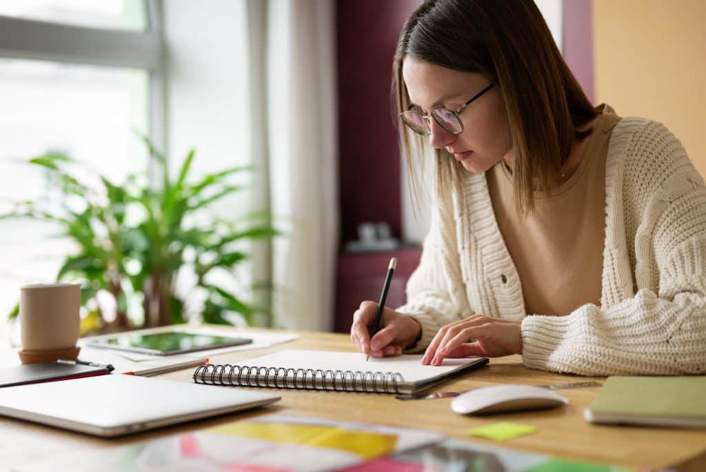 Pessoa usando óculos escrevendo em um caderno em uma mesa, representando o estudo e a dedicação à escrita.