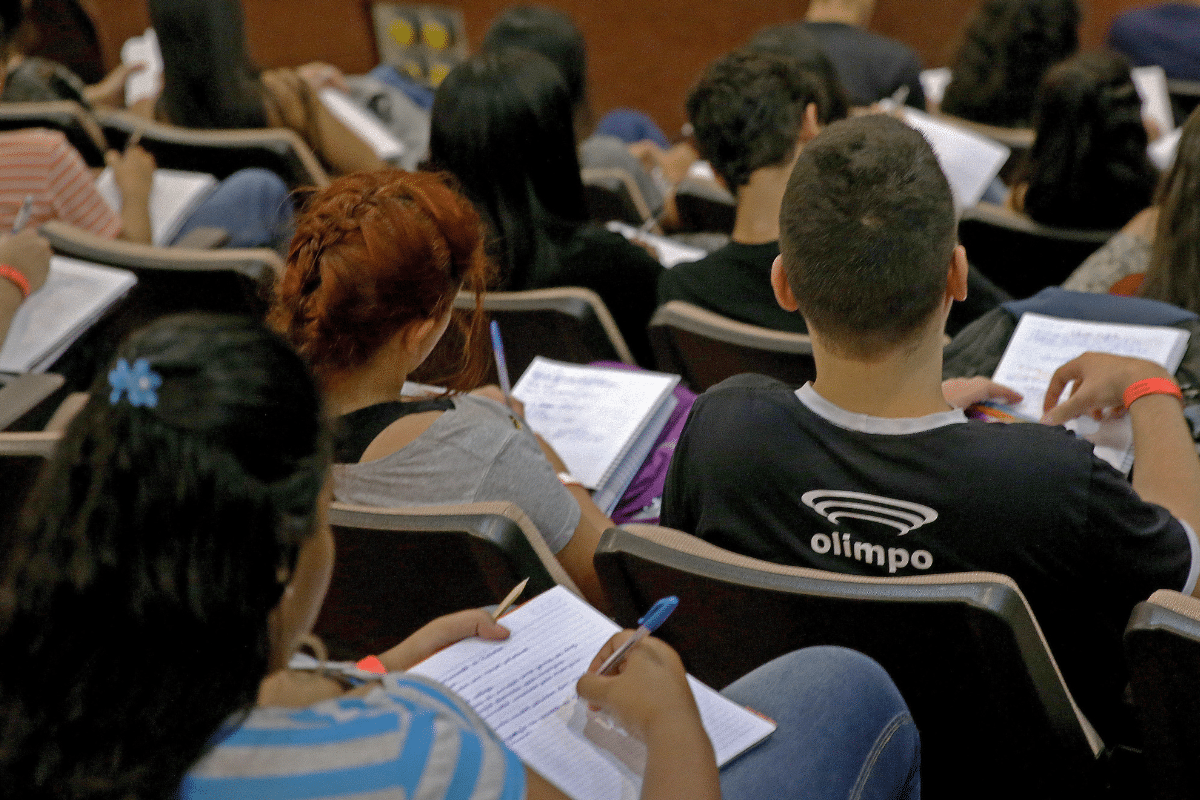 Jovens em auditório acadêmico anotando em cadernos durante aula