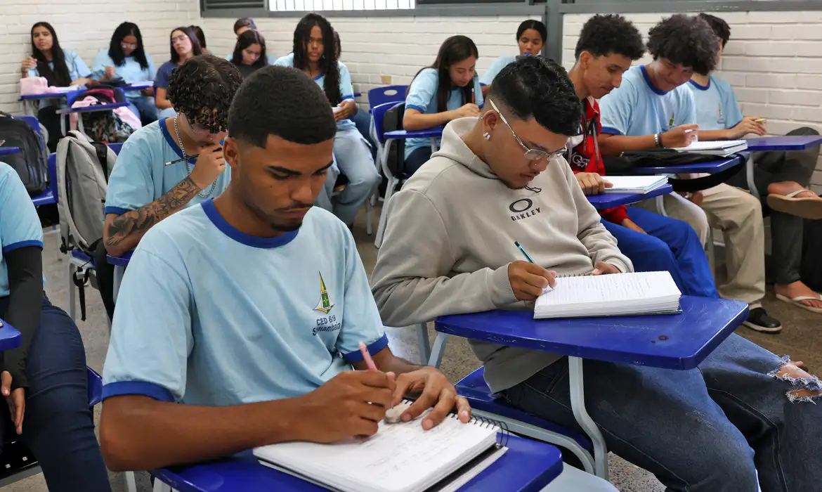  Jovens estudantes em sala de aula escrevendo em cadernos, com uniformes azuis e ambiente escolar organizado