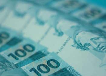 Fundo azul claro com várias notas de 100 reais espalhadas. A imagem está com foco em uma das notas, que mostra a efígie da República e o número "100" em destaque.