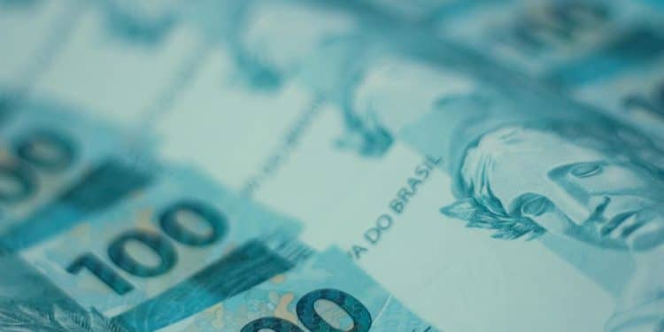 Fundo azul claro com várias notas de 100 reais espalhadas. A imagem está com foco em uma das notas, que mostra a efígie da República e o número "100" em destaque.