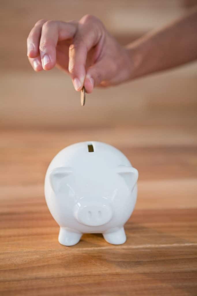 Mão depositando uma moeda em um cofrinho de porquinho branco.