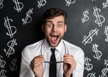 Homem de camisa branca e gravata preta surpreso segurando óculos com fundo de quadro negro e símbolos de dólar desenhados.