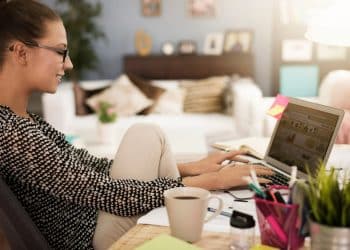 Mulher sorridente usando óculos digitando no laptop enquanto senta com uma perna no braço da cadeira e uma xícara de café na mesa.