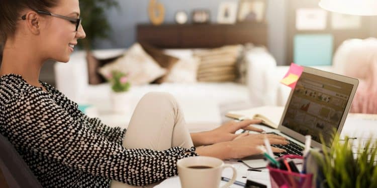 Mulher sorridente usando óculos digitando no laptop enquanto senta com uma perna no braço da cadeira e uma xícara de café na mesa.