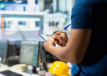 Profissional técnico fazendo anotações em ambiente industrial com equipamentos ao fundo