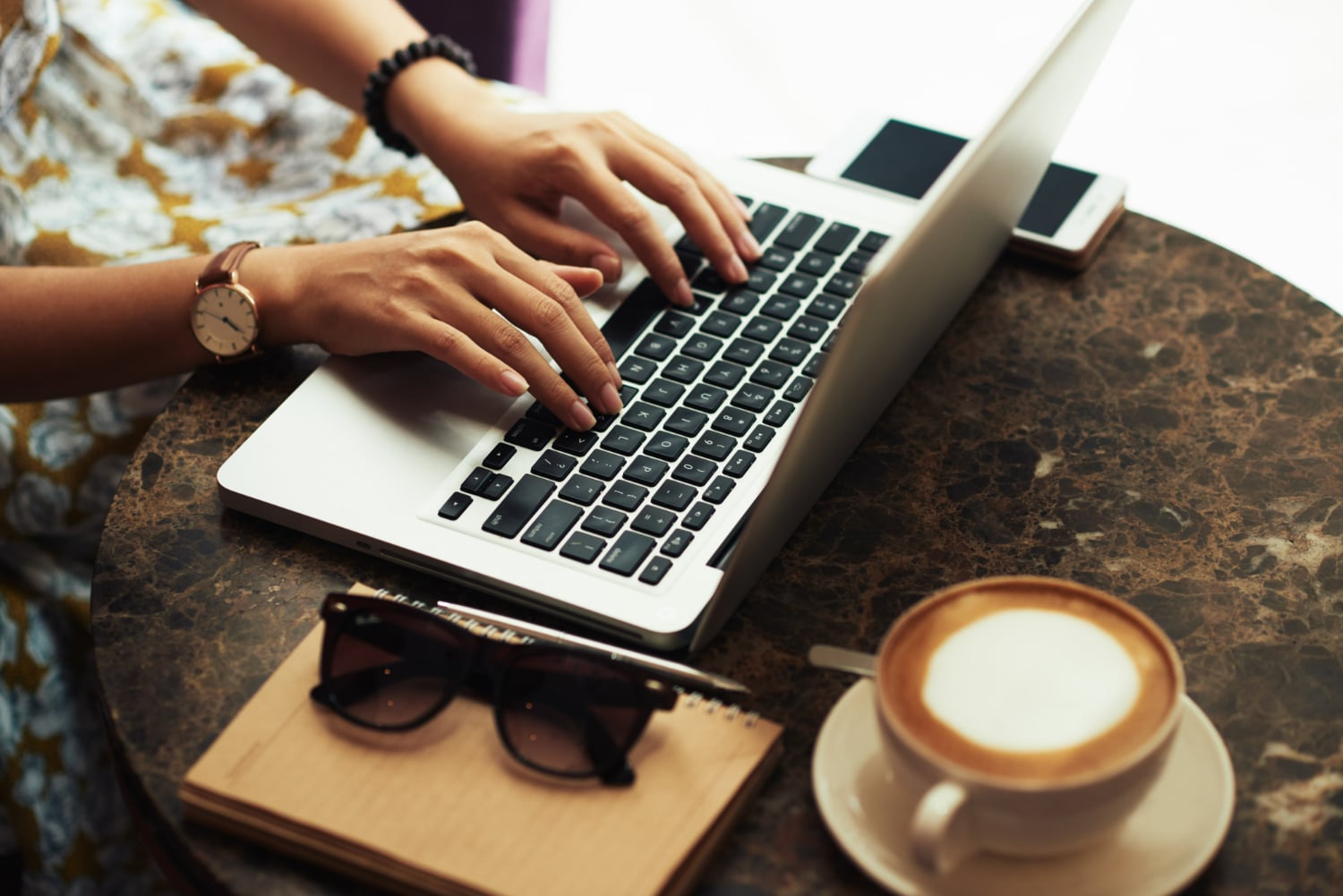 Pessoa digitando no laptop em mesa de pedra com caderno, óculos, celular e xícara de café.