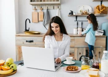 Mulher sorrindo usando notebook na mesa da cozinha com café e comida, criança ao fundo mexendo em objetos.