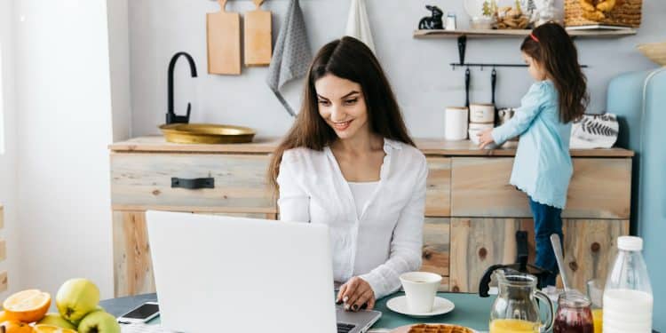 Mulher sorrindo usando notebook na mesa da cozinha com café e comida, criança ao fundo mexendo em objetos.