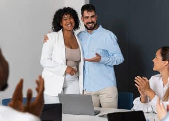 Duas pessoas sorridentes se abraçando e sendo aplaudidas por colegas em reunião profissional.