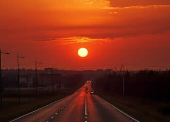 Estrada reta com carros e postes sob pôr do sol intenso com céu laranja e vermelho.