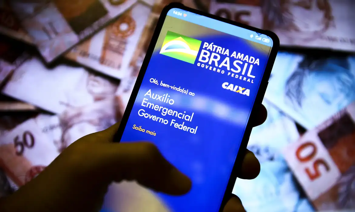 Tela de celular exibindo o aplicativo do Auxílio Emergencial Caixa e notas de R$ 50 no fundo, ilustrando a distribuição do benefício e a polêmica sobre quem não precisa devolver.
