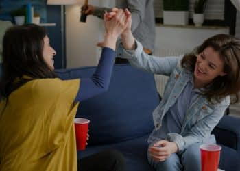 Duas amigas sorridentes brindando com copos vermelhos sentadas em sofá azul.