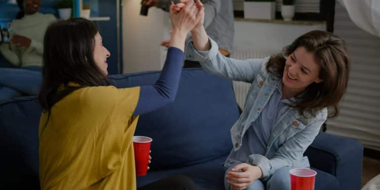 Duas amigas sorridentes brindando com copos vermelhos sentadas em sofá azul.