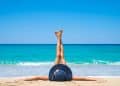 Pessoa deitada na areia da praia com chapéu azul e pernas esticadas para cima na frente do mar azul.
