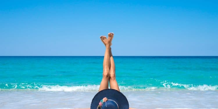 Pessoa deitada na areia da praia com chapéu azul e pernas esticadas para cima na frente do mar azul.
