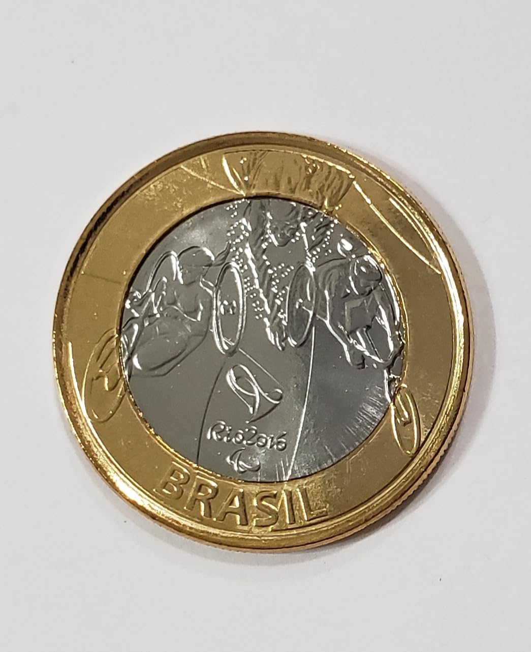 Imagem de alta proximidade da moeda de 1 Real comemorativa do Paratriatlo (Rio 2016), destacando a figura do atleta em cadeira adaptada, o símbolo Paralímpico e a palavra "BRASIL" na borda inferior.