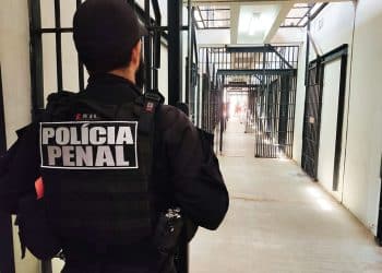 Agente da Polícia Penal de costas em uniforme operacional observando corredor de celas no sistema penitenciário