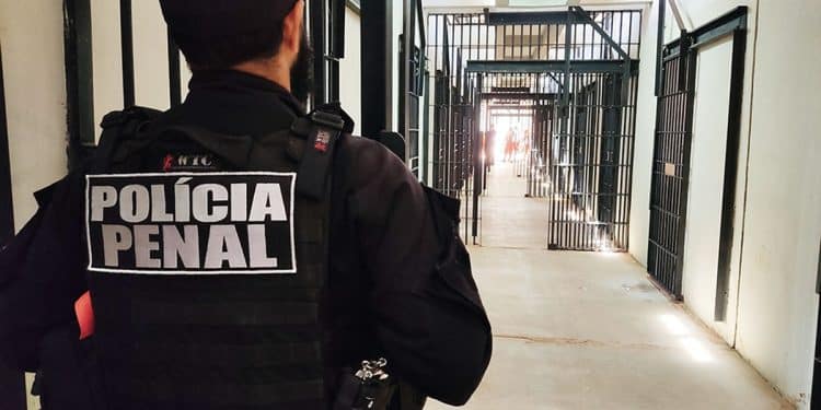 Agente da Polícia Penal de costas em uniforme operacional observando corredor de celas no sistema penitenciário