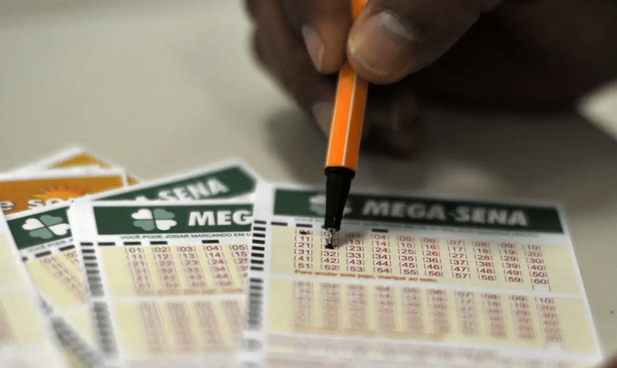 Mão marcando números em volante da Mega-Sena com caneta laranja sobre cartões de apostas da loteria"