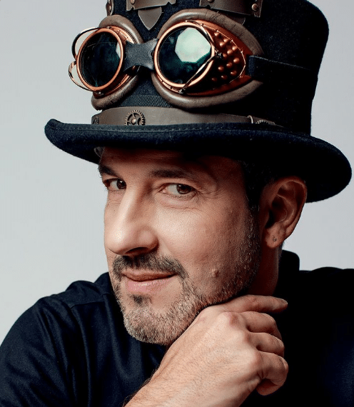 Homem sorridente usando chapéu preto estilo steampunk com óculos metálicos e detalhes em cobre
