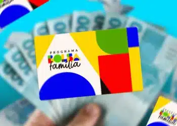 Mão segurando cartão colorido do Programa Bolsa Família com várias notas de dinheiro azul em fundo desfocado