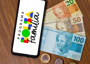 Celular com logo do Programa Bolsa Família e cédulas e moedas de real sobre mesa de madeira.