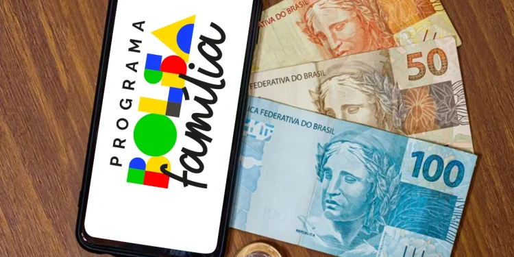 Celular com logo do Programa Bolsa Família e cédulas e moedas de real sobre mesa de madeira.
