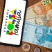 Celular com logo do Programa Bolsa Família e cédulas e moedas de real sobre mesa de madeira.