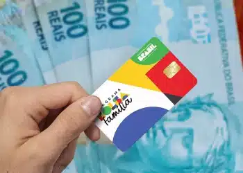 Cartão do Programa Bolsa Família em destaque, segurado por uma mão, com notas de 100 reais ao fundo, ilustrando o início dos pagamentos de outubro.