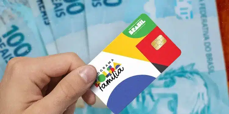 Cartão do Programa Bolsa Família em destaque, segurado por uma mão, com notas de 100 reais ao fundo, ilustrando o início dos pagamentos de outubro.
