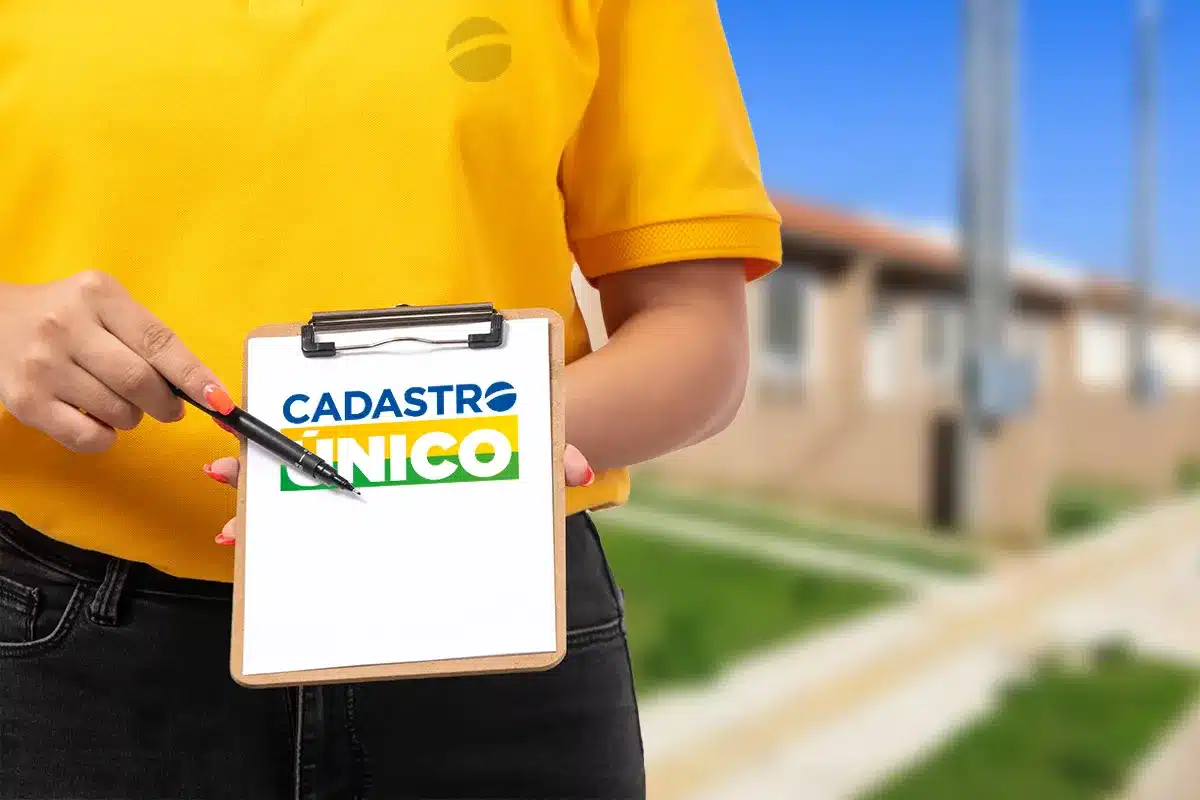 Pessoa vestindo camiseta amarela segurando prancheta com logo do Cadastro Único em área residencial