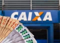 Cédulas de reais em diferentes valores em destaque com a fachada da agência Caixa desfocada ao fundo.