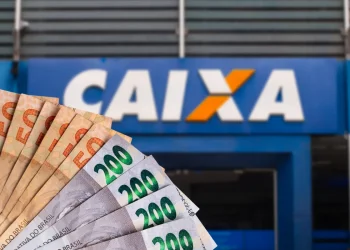 Cédulas de reais em diferentes valores em destaque com a fachada da agência Caixa desfocada ao fundo.