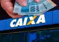 Mão segurando cédulas de real sobre fachada da Caixa Econômica Federal em dia de saque.