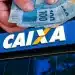 Mão segurando cédulas de real sobre fachada da Caixa Econômica Federal em dia de saque.