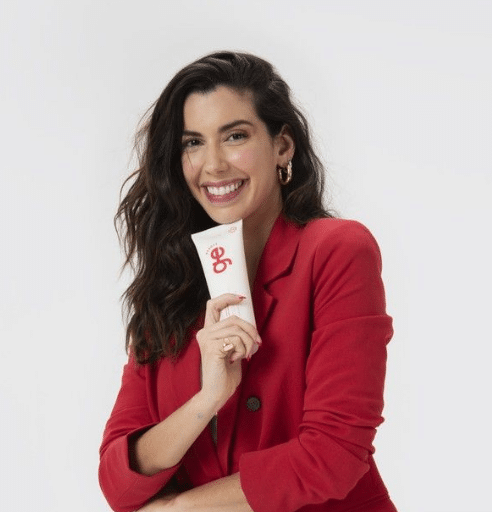 Mulher com cabelos castanhos, sorriso largo, veste blazer vermelho e segura creme facial egê