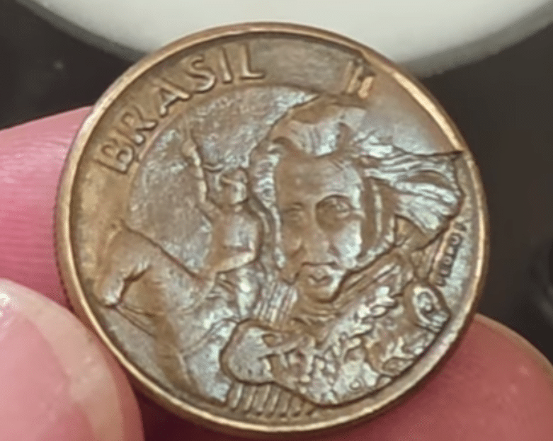 Moeda de 1 Real do Judô (Rio 2016) com um visível erro de cunhagem (trinca ou falha na matriz). A imagem destaca a importância dos defeitos de fabricação na numismática para aumentar o valor da peça.