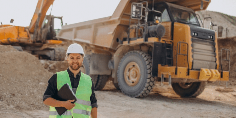 Técnico em Mineração com capacete e colete refletivo em canteiro de obras, em frente a máquinas pesadas.