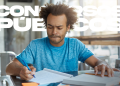 Jovem vestindo camiseta azul estudando para concursos em mesa com livros e caderno.