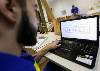 Homem com barba usando camiseta azul trabalha em notebook Samsung com software aberto em ambiente de escritório