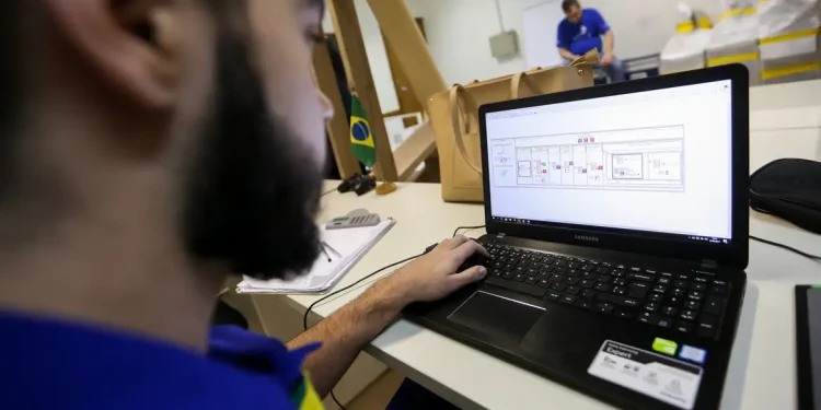 Homem com barba usando camiseta azul trabalha em notebook Samsung com software aberto em ambiente de escritório