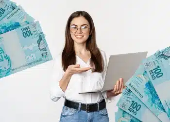 Imagem de uma mulher com laptop e cédulas de 100 reais, simbolizando o reajuste do salário mínimo em 2026