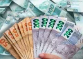 Mão segura leque de notas de 50, 100 e 200 reais sobre uma pilha de dinheiro, ilustrando meta de R$ 100 mil