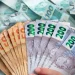 Mão segura leque de notas de 50, 100 e 200 reais sobre uma pilha de dinheiro, ilustrando meta de R$ 100 mil