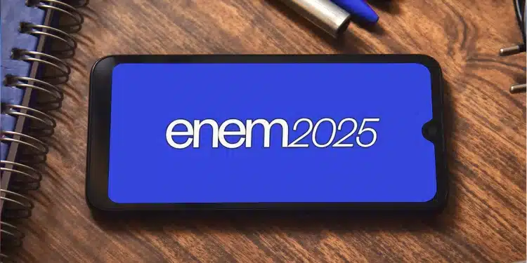Tela de celular exibindo logo ENEM 2025 em fundo azul sobre mesa com caderno e canetas.