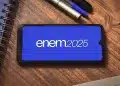 Celular na mesa exibindo o logo ENEM 2025 em fundo azul, com caderno e caneta ao lado