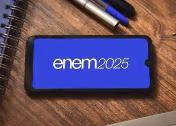 Celular na mesa exibindo o logo ENEM 2025 em fundo azul, com caderno e caneta ao lado
